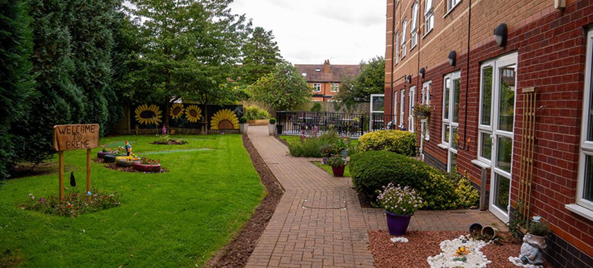 A tidy paved outdoor walkway joining the property to the outdoor grassed area. The walkways are decorated with a mixture of shrubbery and potted plants.