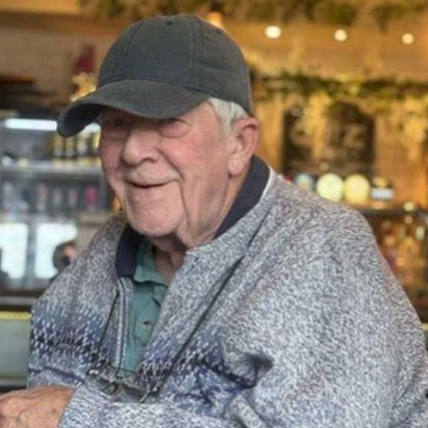 An elderly man seated at a table, holding a cup of coffee and smiling softly.