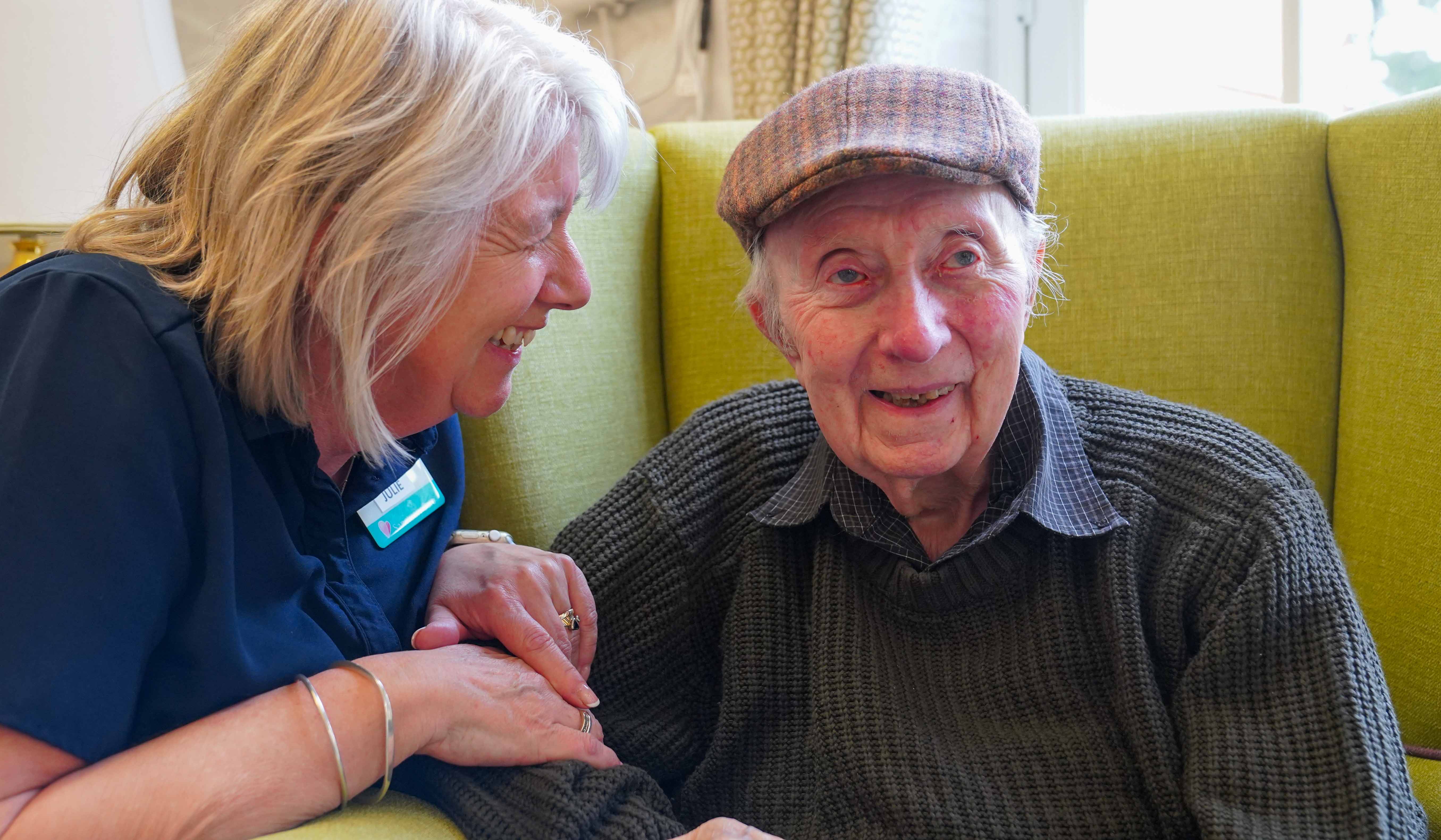 A male resident sat in a comfortable yellow arm chair with a member of staff smiling with him and comforting him