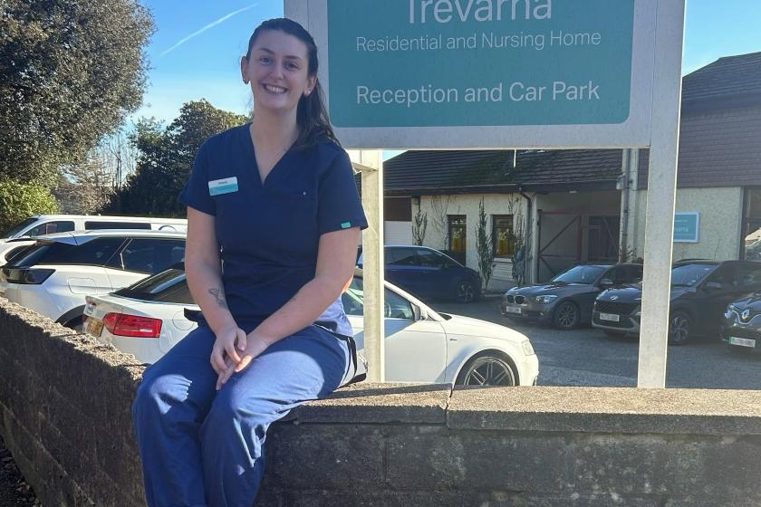 A young woman wearing blue medical scrubs sitting on a brick wall in front of a sign that reads 'Trevarna Residential and Nursing Home'