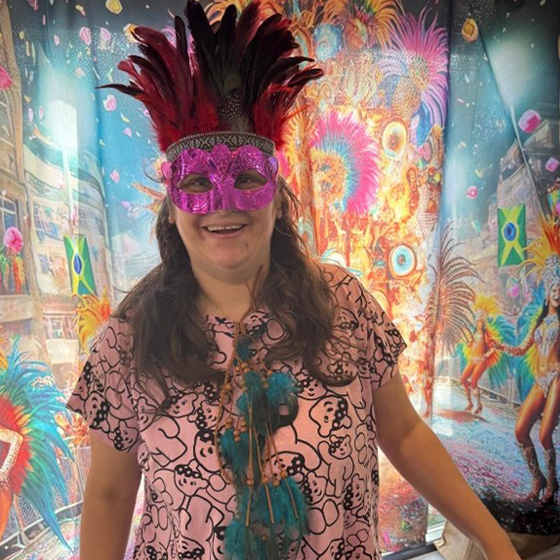 A person in a pink shirt and red feathered headdress stands before a vibrant carnival backdrop with Brazilian flags.