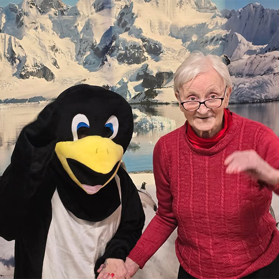 A person in a penguin costume stands beside an elderly person in a red sweater, with a snowy mountain and lake scene in the background.