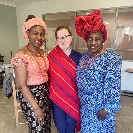 Three people standing indside, two wear traditional African attire with headwraps, and one wears a red scarf and blue outfit.