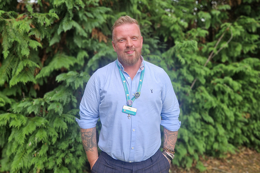 Sanctuary Care Property Manager Tony Kirkby A man with a beard stands in front of a dense green hedge. He is wearing a light blue button-up shirt and has tattoos visible on his arms. A lanyard with a badge hangs around his neck. The background features lush foliage, creating a natural setting.