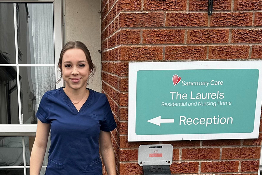A person in navy blue scrubs standing outside a brick residential and nursing home beside a sign reading ‘Sanctuary Care – The Laurels – Residential and Nursing Home"