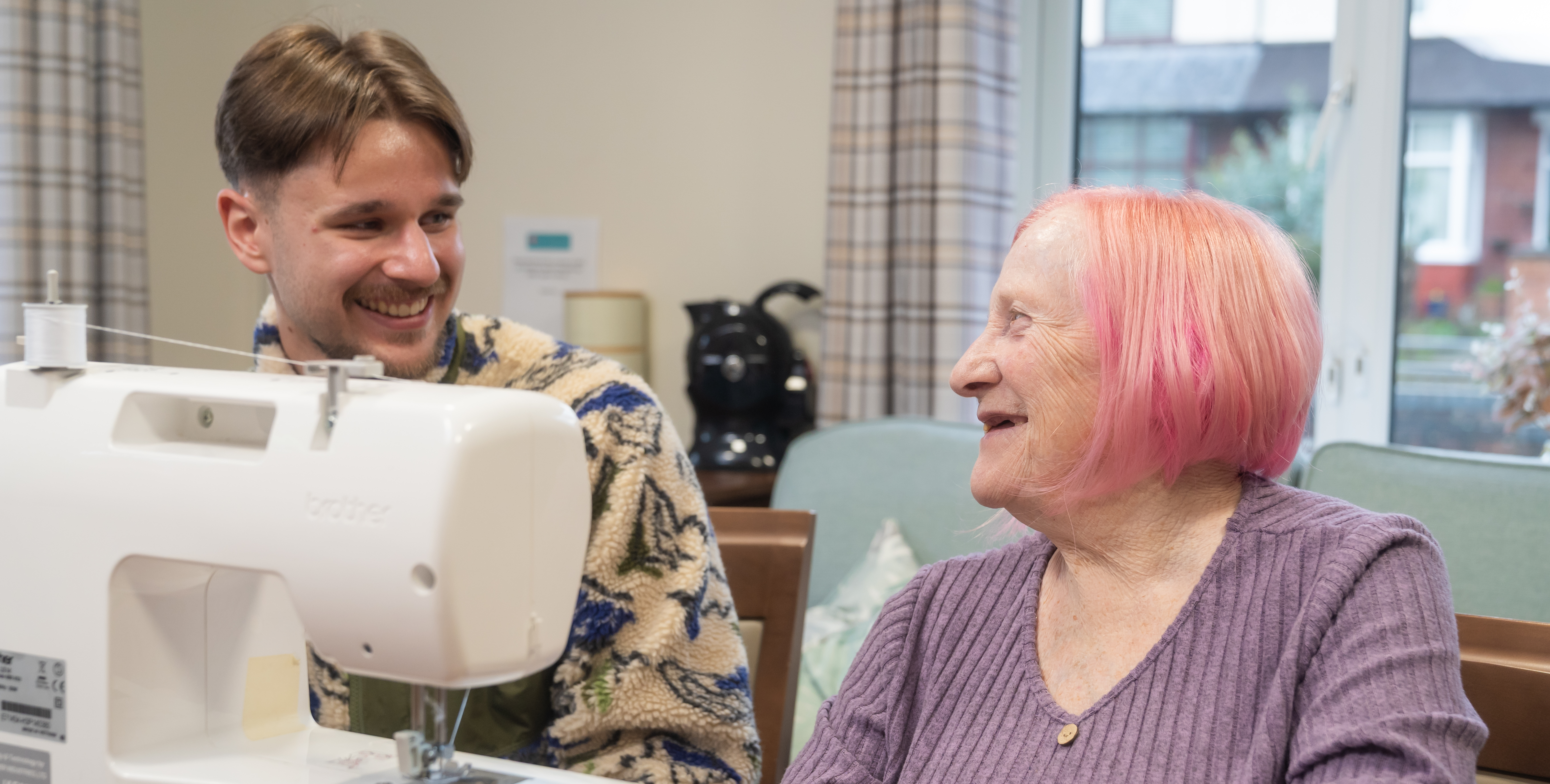 Two people smiling while sitting indoors beside a white sewing machine, one older with pink hair and one younger with short brown hair, in a cozy room with a window and coffee machine in the background.