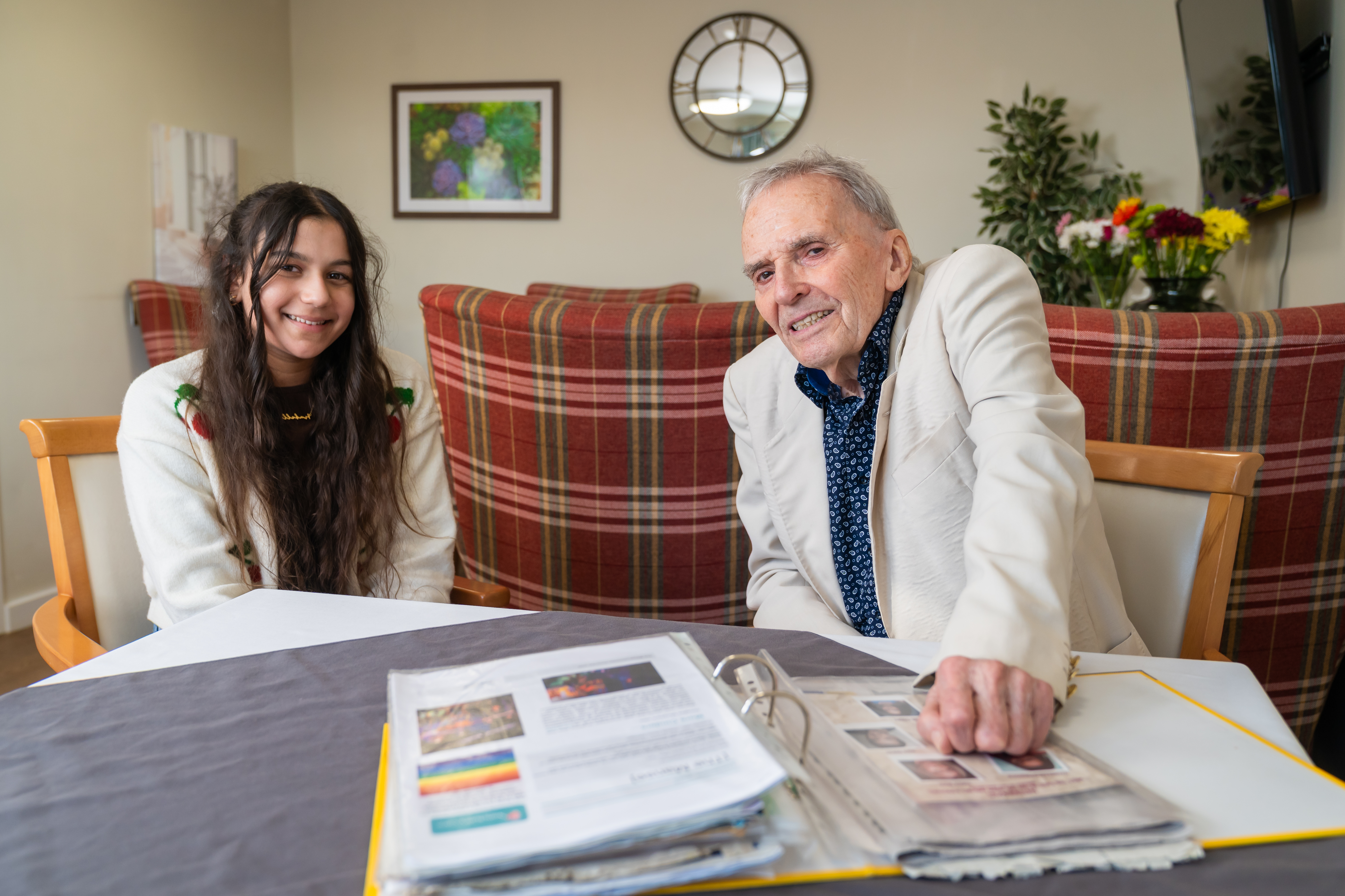 Two people sitting at a table in a care home lounge, looking through a folder, with plaid furniture, wall art, and flowers in the background