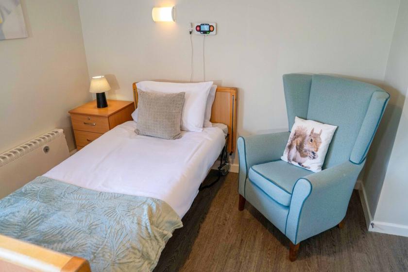 A clean minimal bedroom with a medical single bed with an oversized blue arm chair.