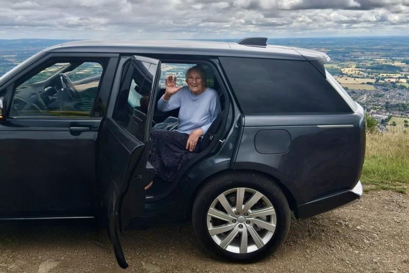 Person seated in the back of a black SUV with the rear door open, waving on a dirt road with scenic landscape and cloudy sky in the background