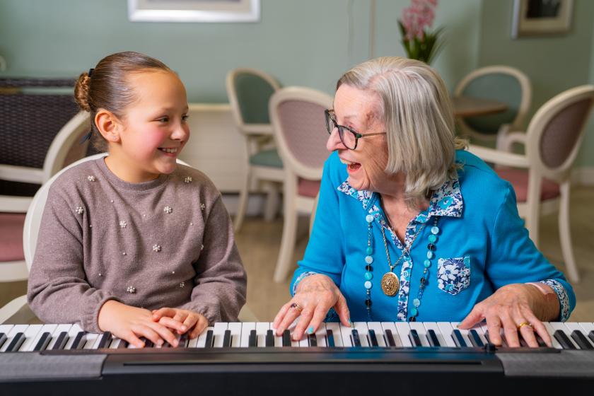 Young girl and resident Margaret smiling together while playing a keyboard piano in a cosy, pastel-coloured room