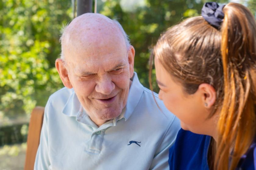 An elderly man with a warm smile is sitting outdoors, engaging in conversation with a young woman.