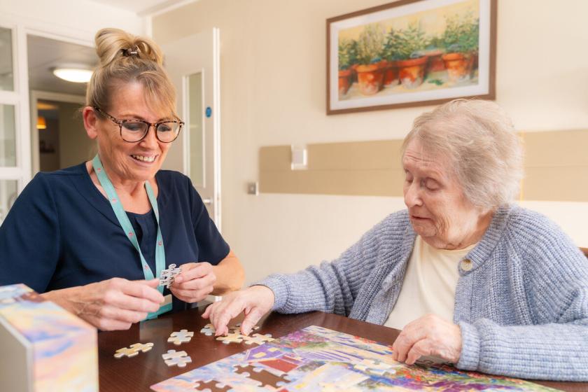 A care worker and resident sat at a table together smiling and doing a puzzle