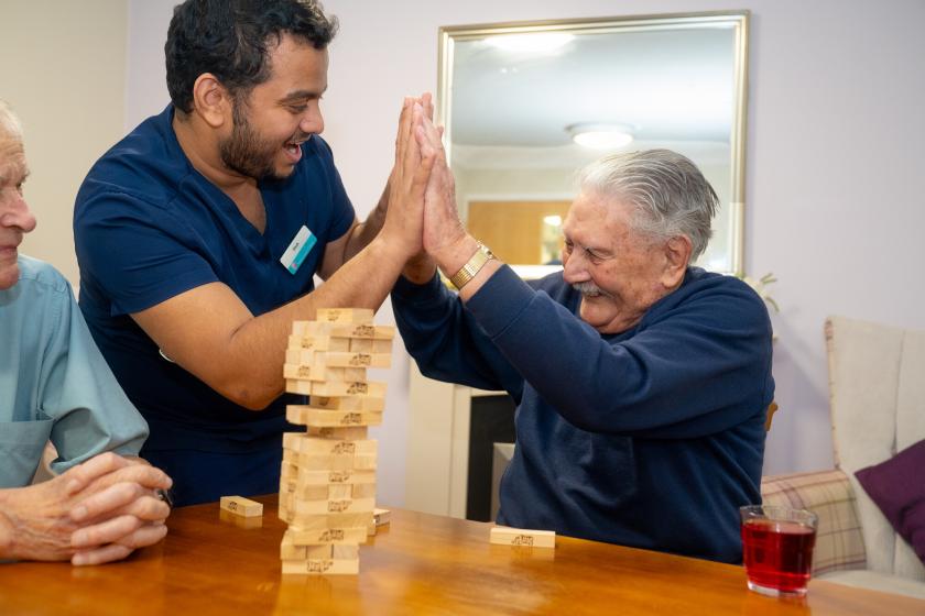 A caregiver and a man are engaged in a playful game of Jenga at a table. The caregiver, wearing a blue uniform, is smiling and raising his hands in excitement as he interacts with the elderly man, who is also smiling and raising his hands in a playful gesture. A partially built Jenga tower is in front of them, and another elderly man sits to the left, observing the game. A glass of red drink is on the table. The setting appears warm and inviting, suggesting a friendly and supportive environment.