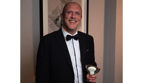 A man in a black tuxedo with a bow tie stands smiling, holding a trophy in one hand. He has short hair and is positioned in front of a framed artwork on the wall. The setting appears to be an elegant indoor space, likely at an awards ceremony or formal event.
