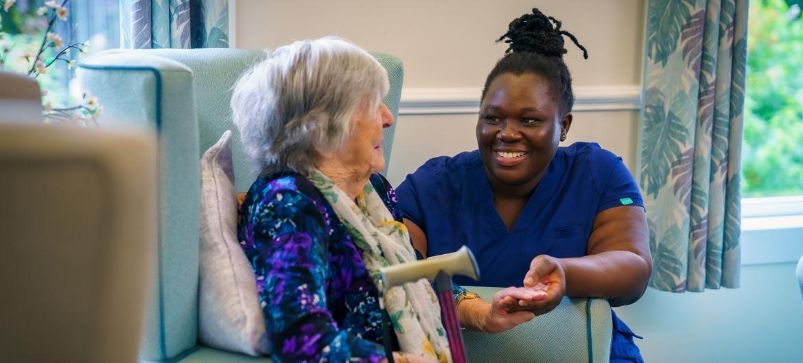 Resident, Betty sat down on a chair looking at Care employee Doreen who is knelt beside her. They are having a conversation and smiling