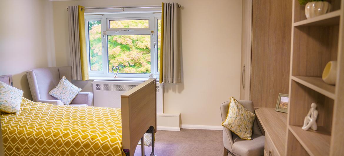 Internal of a bedroom with wooden fittings and yellow bedsheets and curtains. There is a window with a view of the garden on the far wall