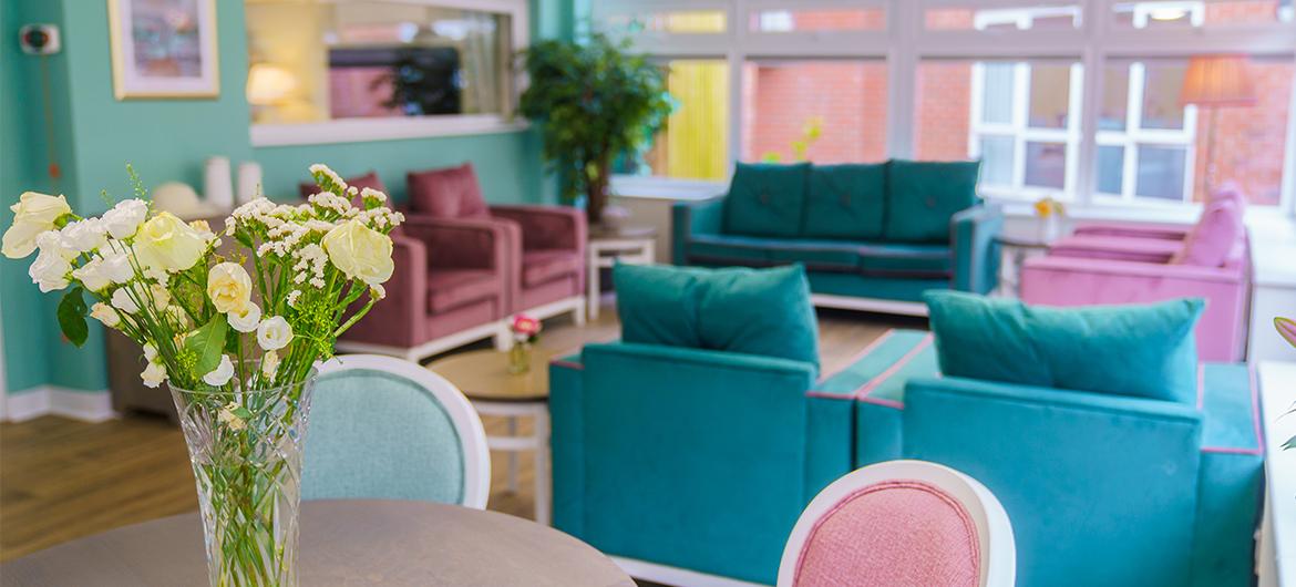 A glass vase holding white and yellow flowers in the foreground with a collection of bright coloured armchairs and sofas in the background