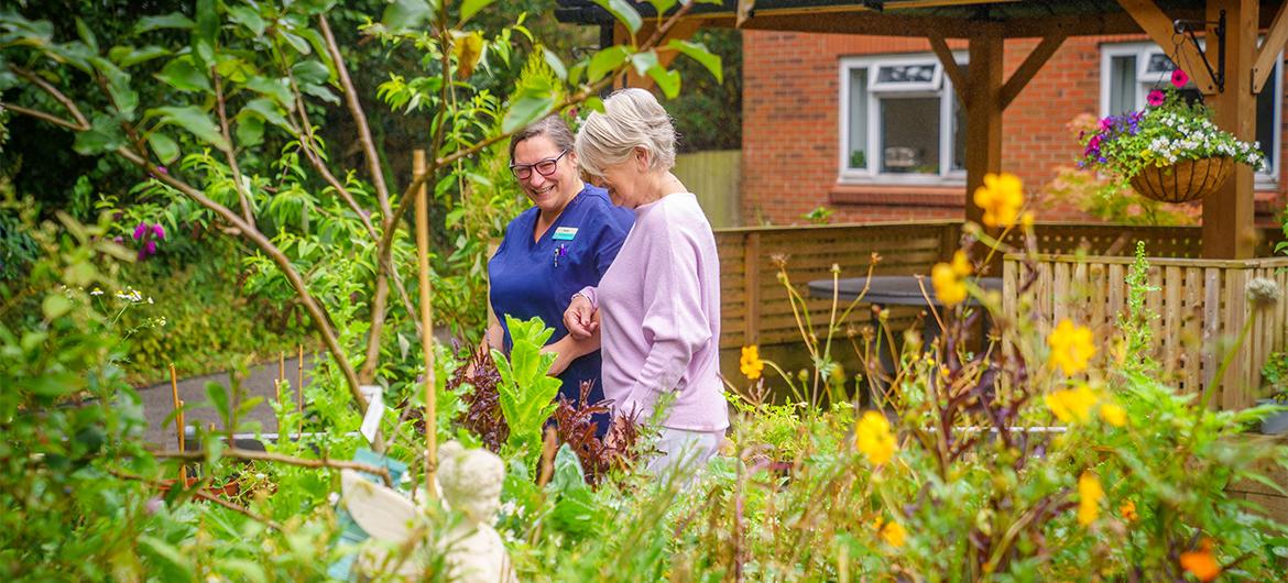 A Sanctuary Care resident and staff member walking through a garden with tall plants as they link arms