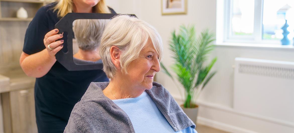 An lady sitting in a chair with a towel around her neck while somebody behind her holds a mirror behind her so she can see the back of her haircut