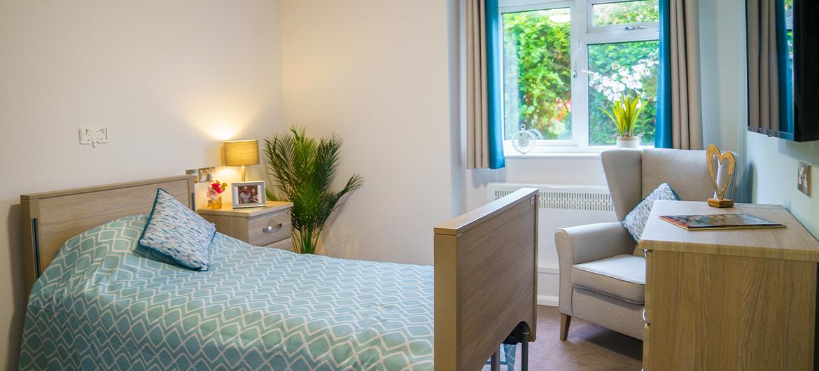 Interior of a bedroom with a single bed with blue bedding, wooden chest of drawers, bedside table, armchair and a window looking into a garden