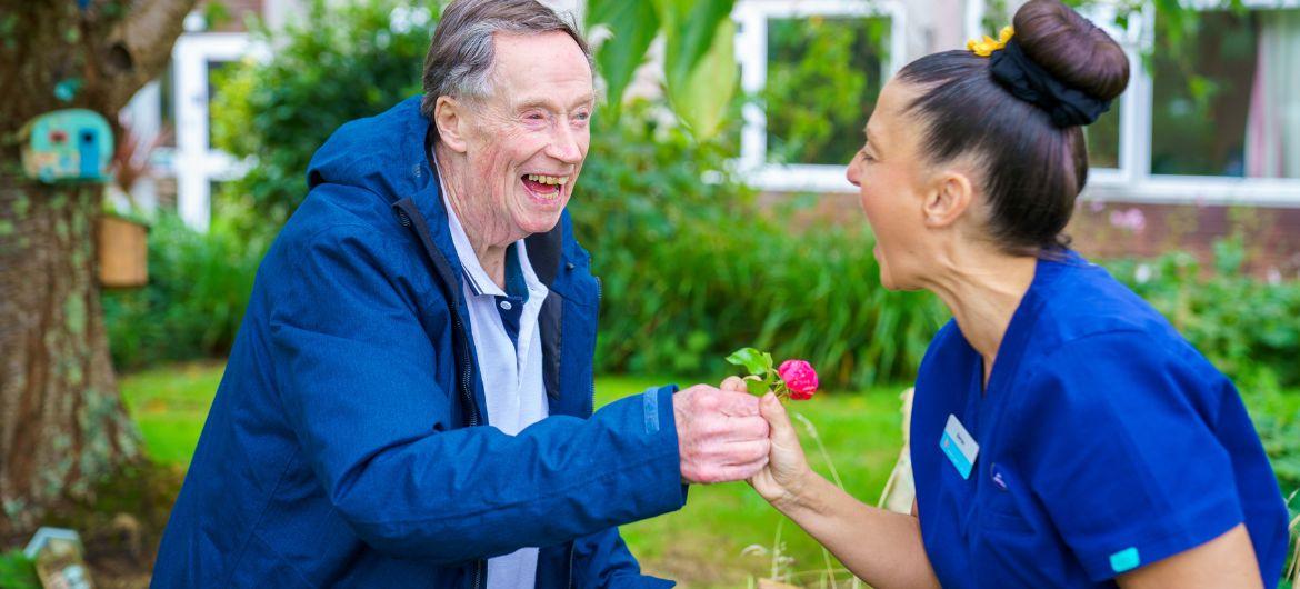 Resident John in the garden with Sanctuary Care Employee Mary. The two are facing eachother and laughing whilst John hands Mary a flower