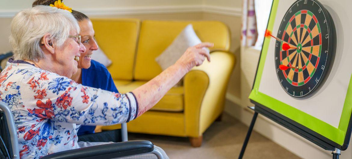 Resident Maureen, sat down playing darts with a Care Employee assisting her