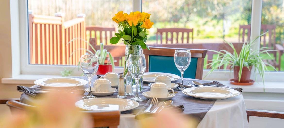 A dinner table with crockery and glassware on with a vase of yellow roses in the middle