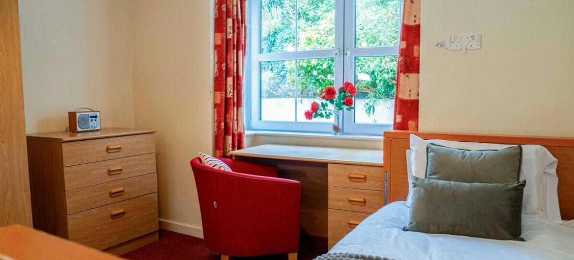 The Trevern, bedroom. A warm and inviting single bedroom, nostalgic touches like a vintage radio, and a comfy armchair by a sunny window with views of greenery.