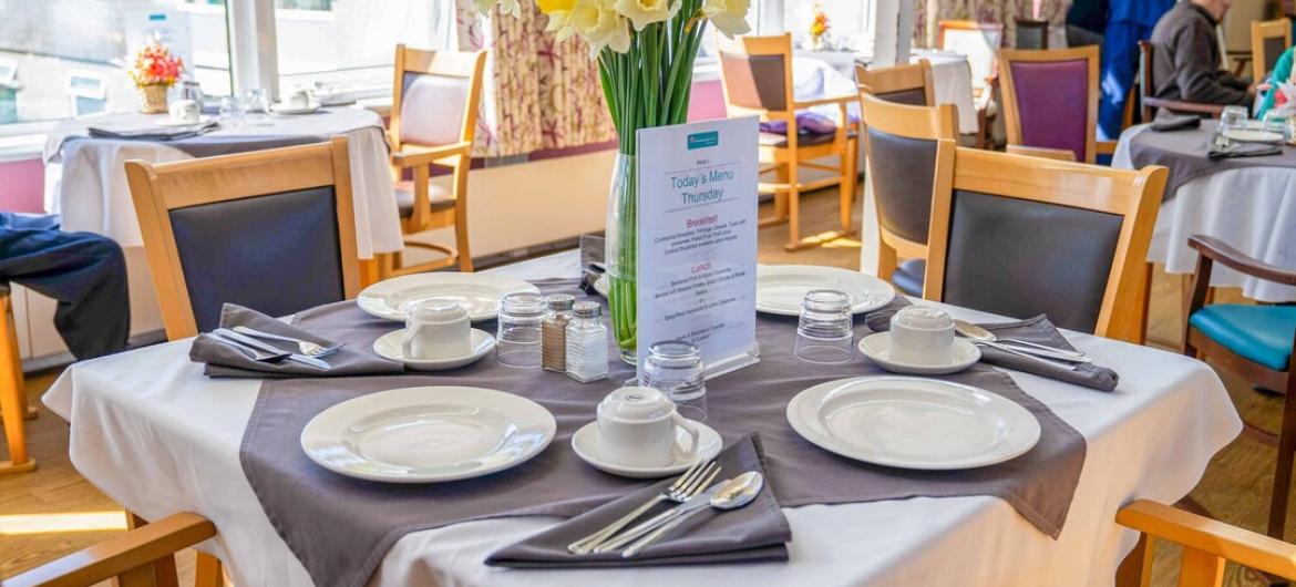 The Woodland, dining room. A bright, welcoming dining area at The Woodland, with neatly set tables, fresh flowers, and large windows letting in natural light. Creating a warm, friendly, and community-focused atmosphere.