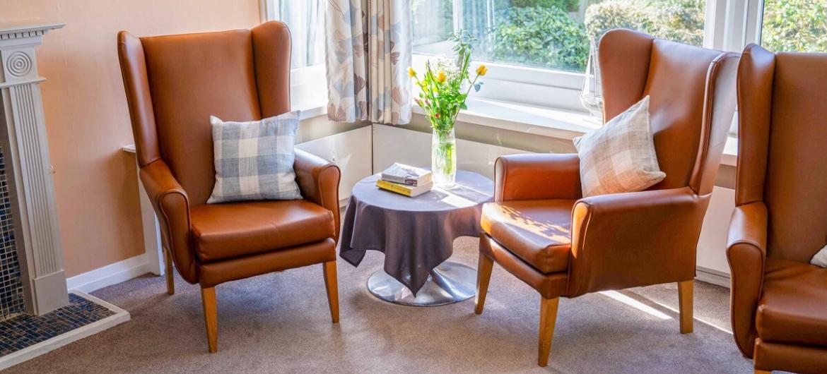 The Woodland, seating area. A bright and cosy sitting area, with comfortable large leather armchairs, soft cushions, and a small table topped with fresh flowers and books. Large windows fill the space with natural light, creating a warm and welcoming environment.