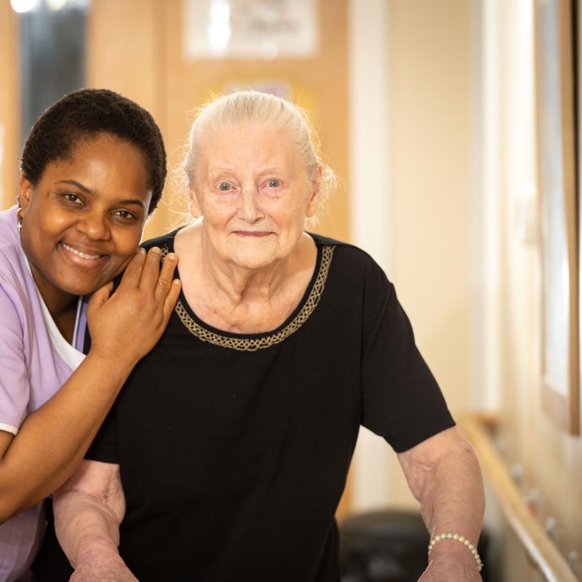 Carer and care home resident stand smiling in corridor 