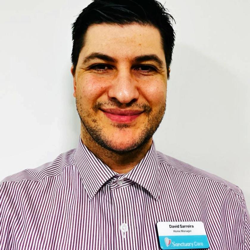 A young man wearing a pinstriped shirt with a Sanctuary Care name badge standing against a white wall