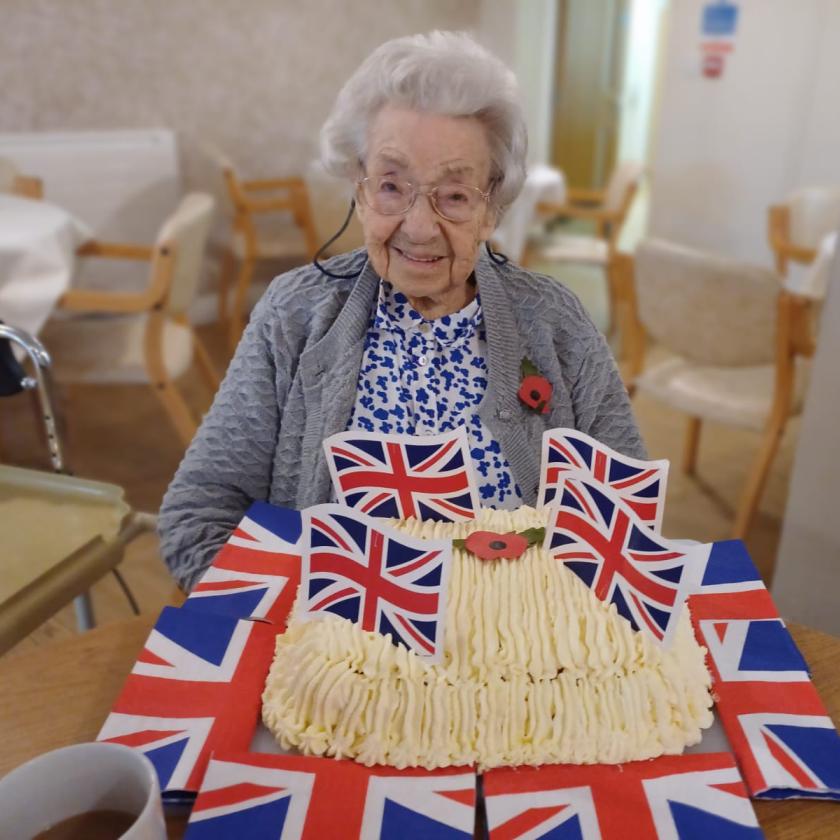 Resident Ruby sat down at a table with a large frosted cake in front of her which is surrounded by union jack flags. She is also wearing a poppy on her cardigan