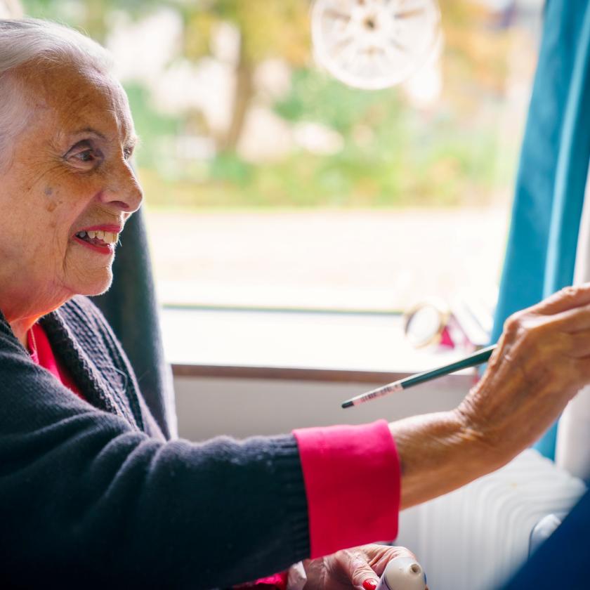 An elderly woman sitting on a sofa and smiling while painting with a paint brush