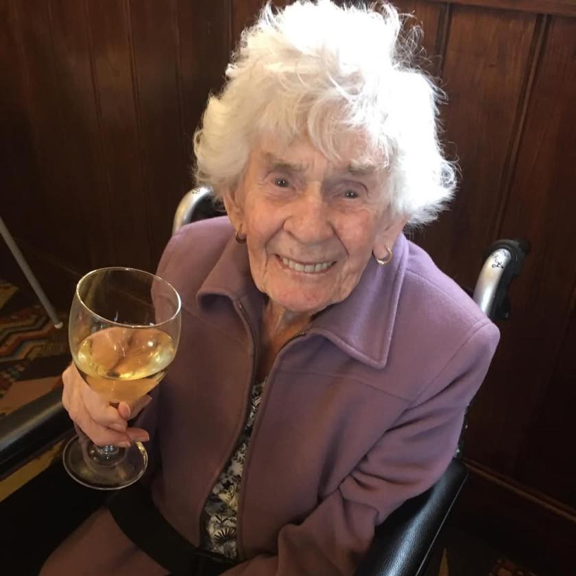 An elderly woman sitting in a wheelchair and smiling while holding a glass of white wine