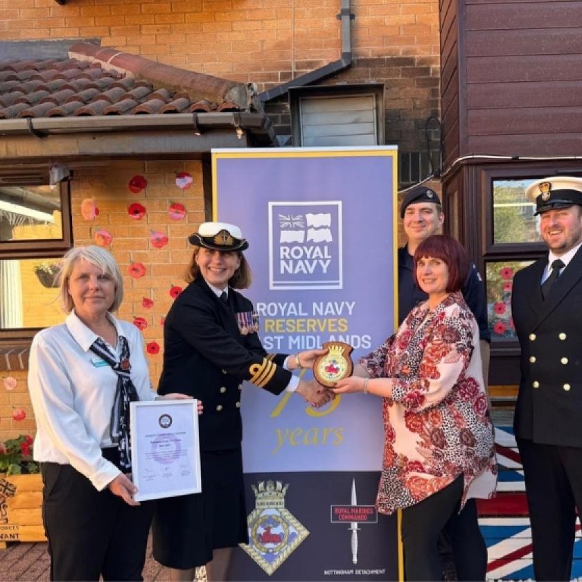 Four individuals standing in front of a banner reading 'ROYAL NAVY RESERVES WEST MIDLANDS.' Two are in Royal Navy uniforms; one holds a certificate, and another hands an object to a person in civilian clothing. A brick building with poppy decorations and a Union Jack flag design is in the background.