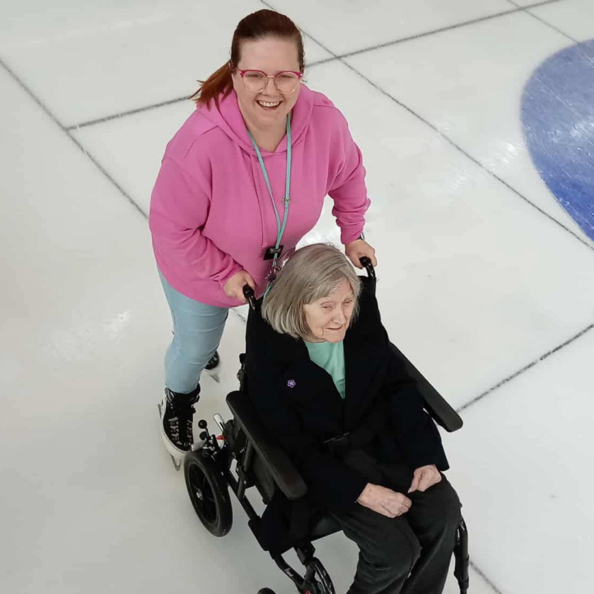 Activities coordinator Kirsty smiles at the camera as she pushes a resident around the ice rink in her wheelchair