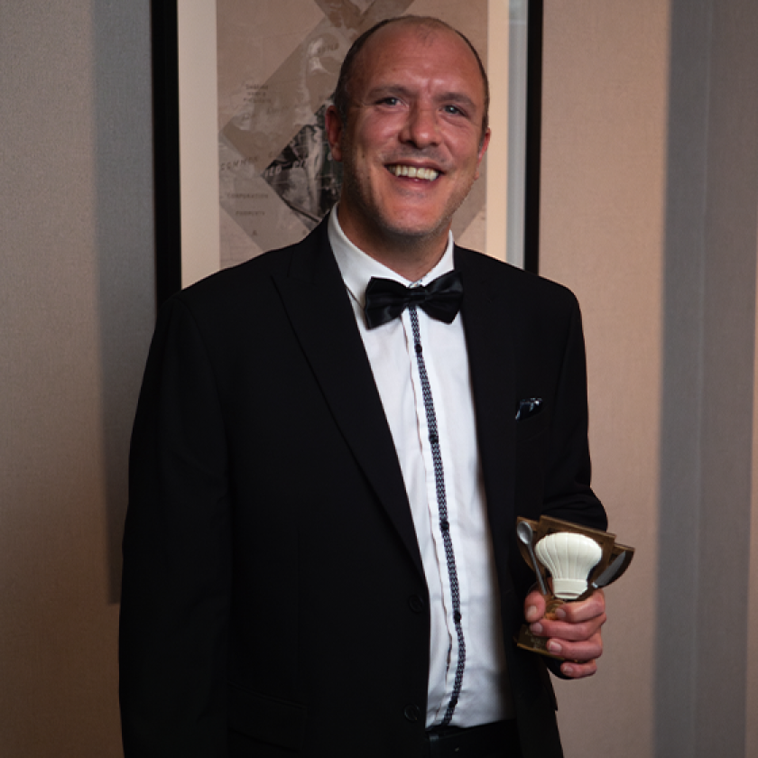 A man in a black tuxedo with a bow tie stands smiling, holding a trophy in one hand. He has short hair and is positioned in front of a framed artwork on the wall. The setting appears to be an elegant indoor space, likely at an awards ceremony or formal event.